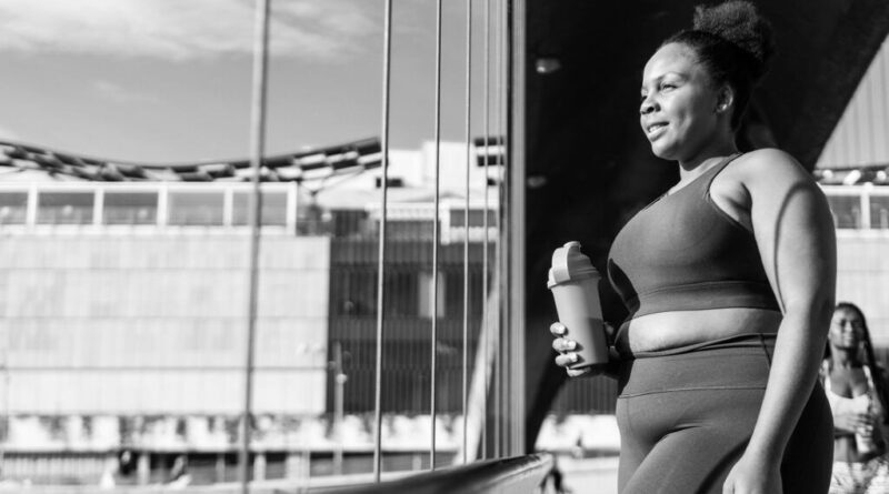 a black woman in gym clothes looks out the window holding a water bottle